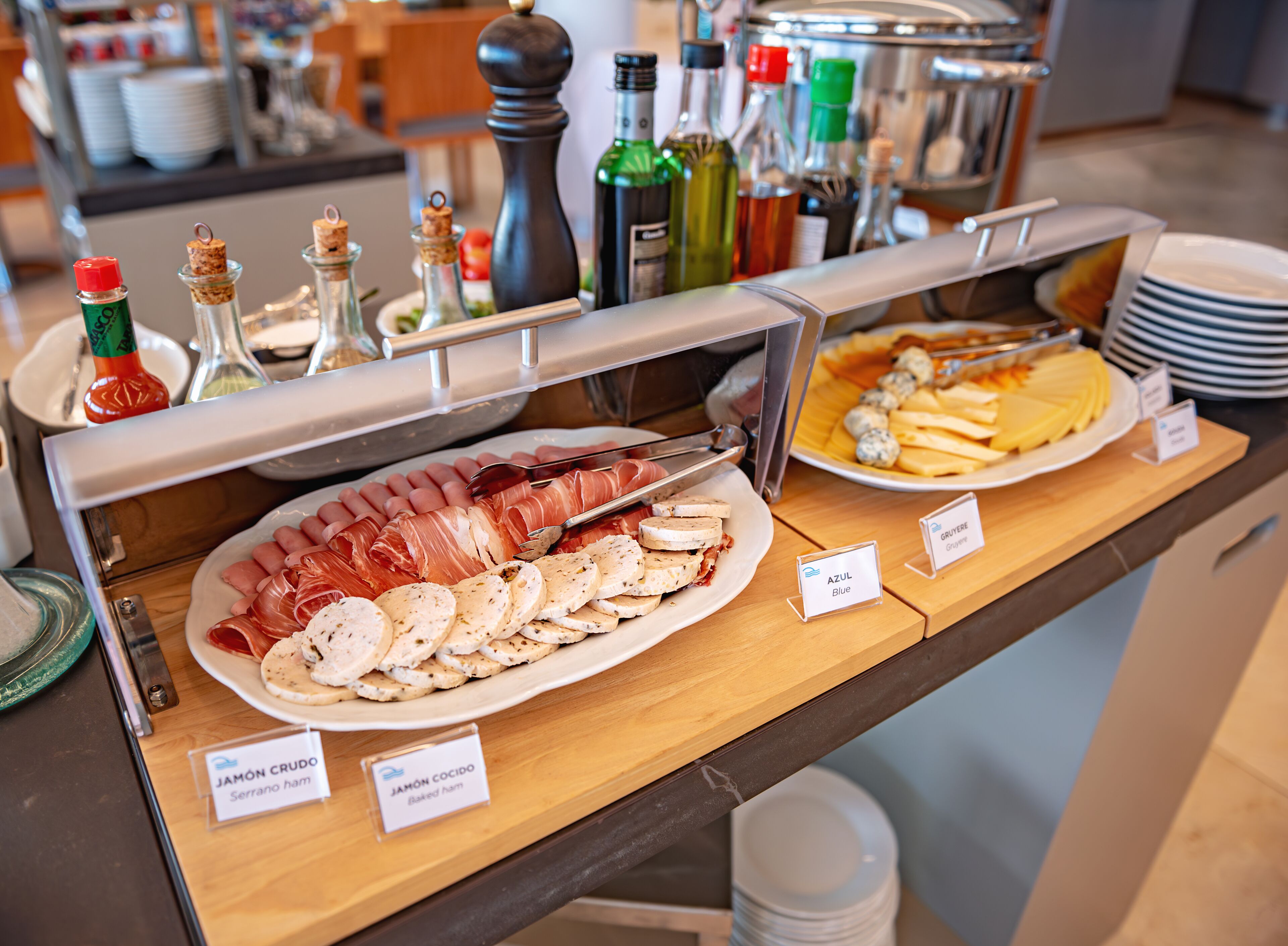 A buffet with various food items including sliced meats and cheeses, labeled with names like "JAMÓN CRUDO" and "AZUL Blue".