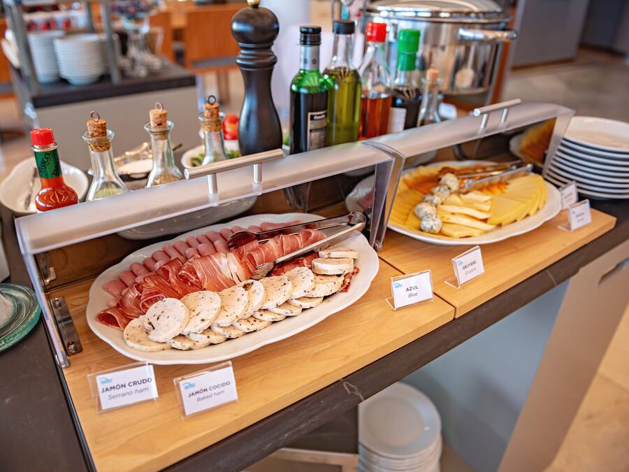 A buffet with various food items including sliced meats and cheeses, labeled with names like "JAMÓN CRUDO" and "AZUL Blue".
