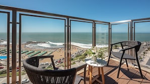 Varanda com vista para a praia e o oceano. Há duas cadeiras e uma pequena mesa com um vaso de flores.