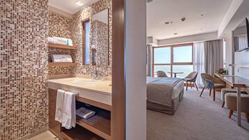 A modern hotel room with a bathroom featuring mosaic tile walls, a large mirror, and a wooden vanity. The room includes a bed, a desk, two chairs, and a view of the sea through large windows.