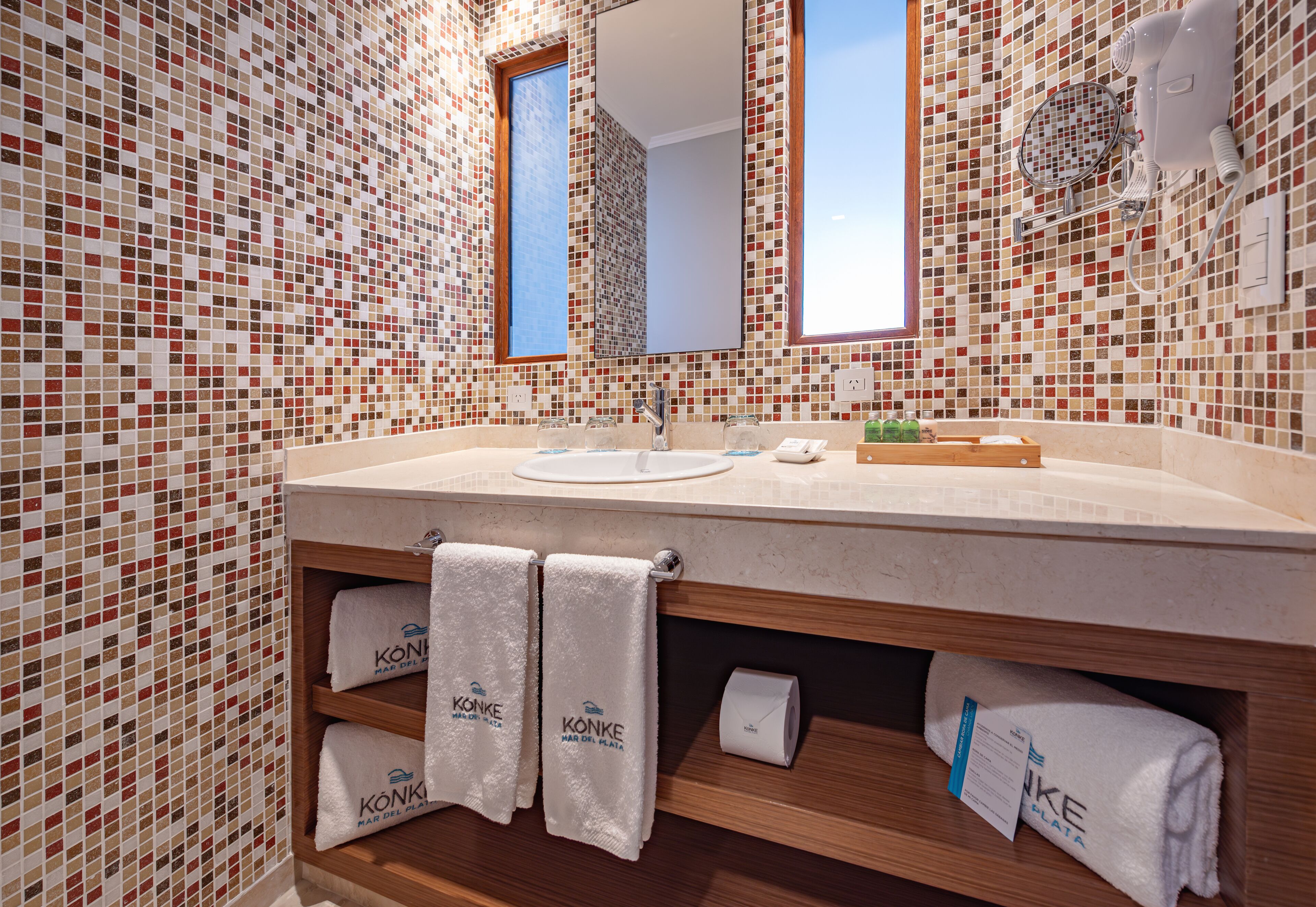 Un baño con paredes de mosaicos, un mueble de tocador de madera, un lavabo, un espejo y toallas con la marca 'KONKE'.