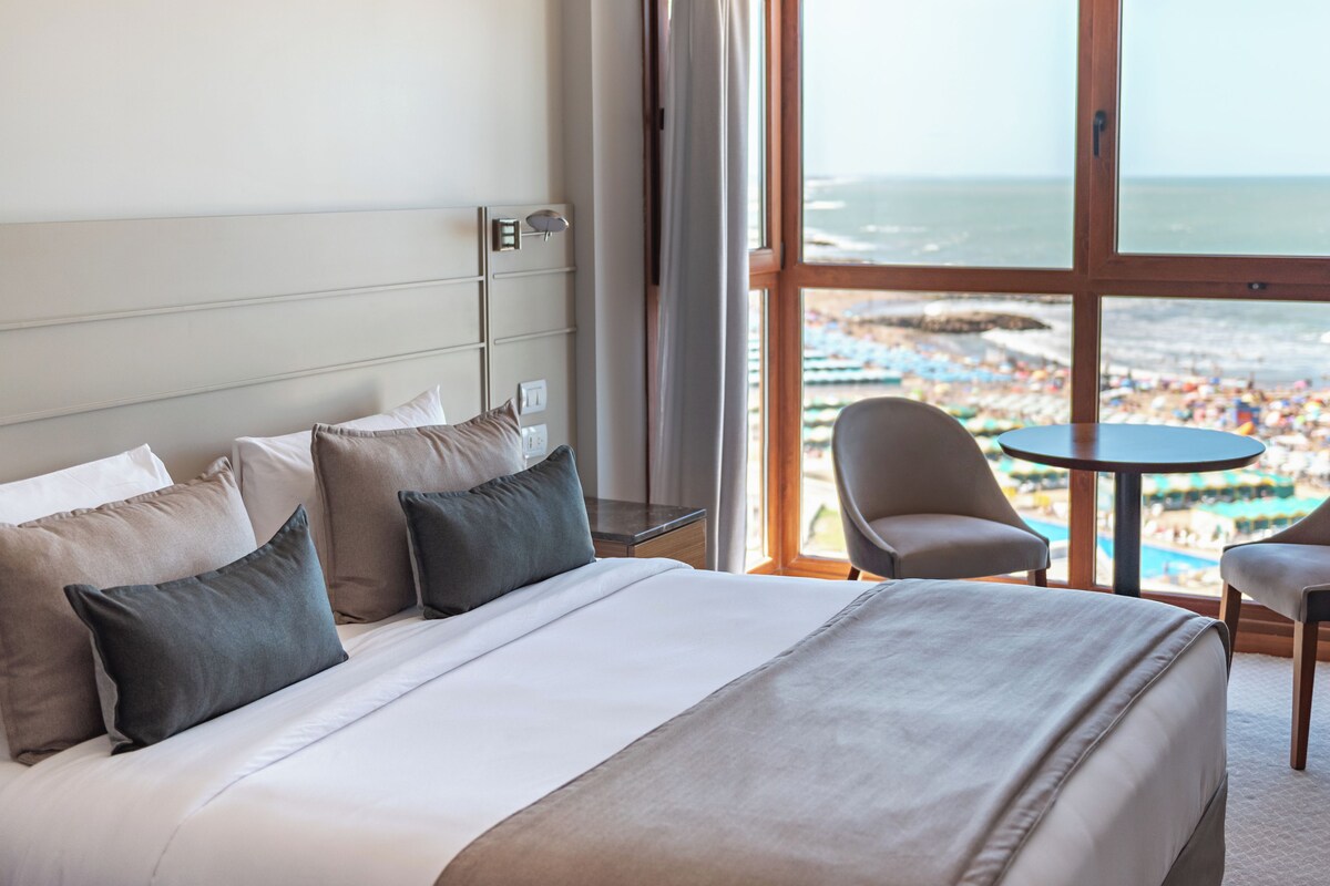 A hotel room with a large bed, two chairs, a small table, and a view of the beach and ocean.