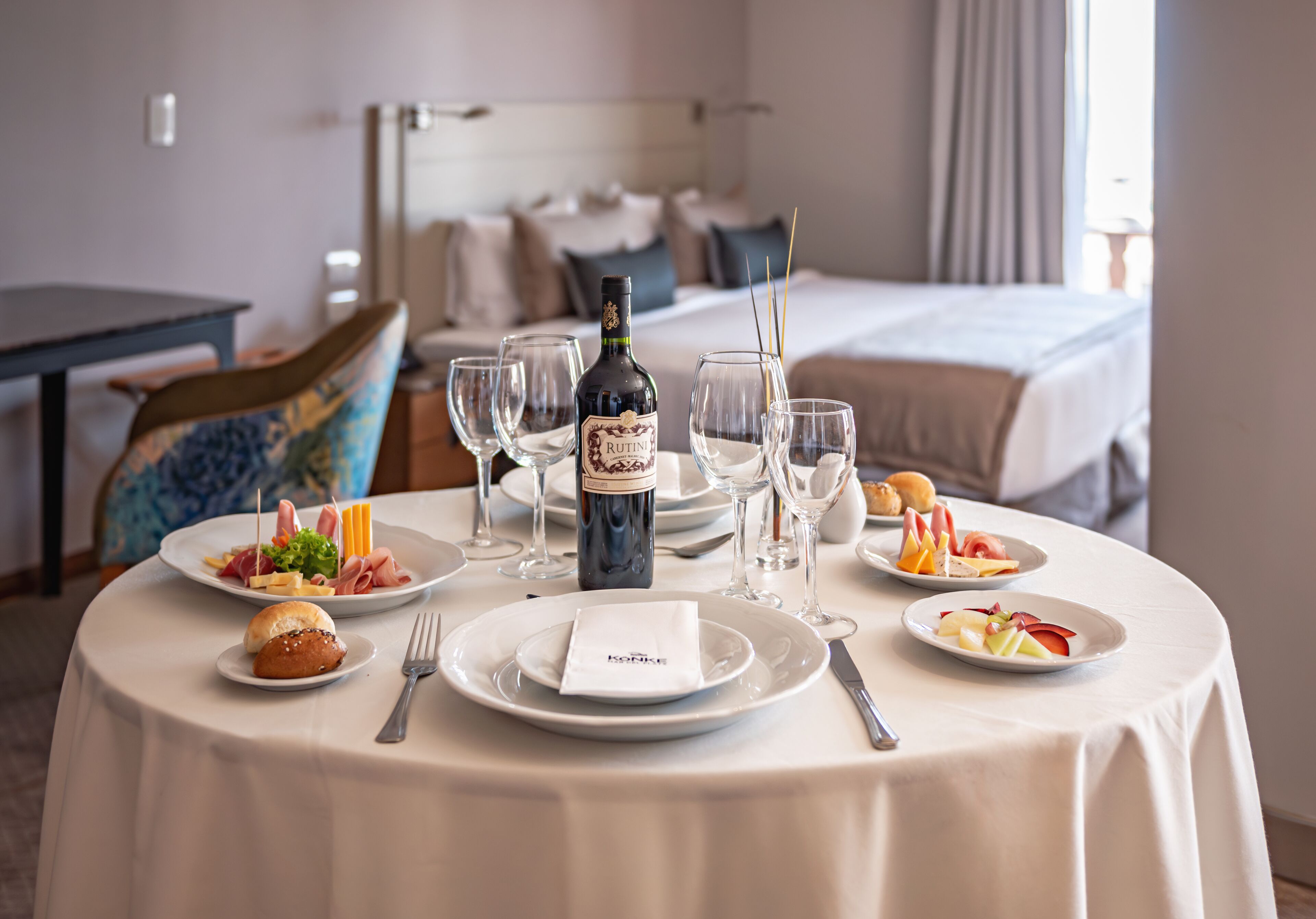 Mesa de comedor de hotel dispuesta con platos, cubiertos, copas y una botella de vino.