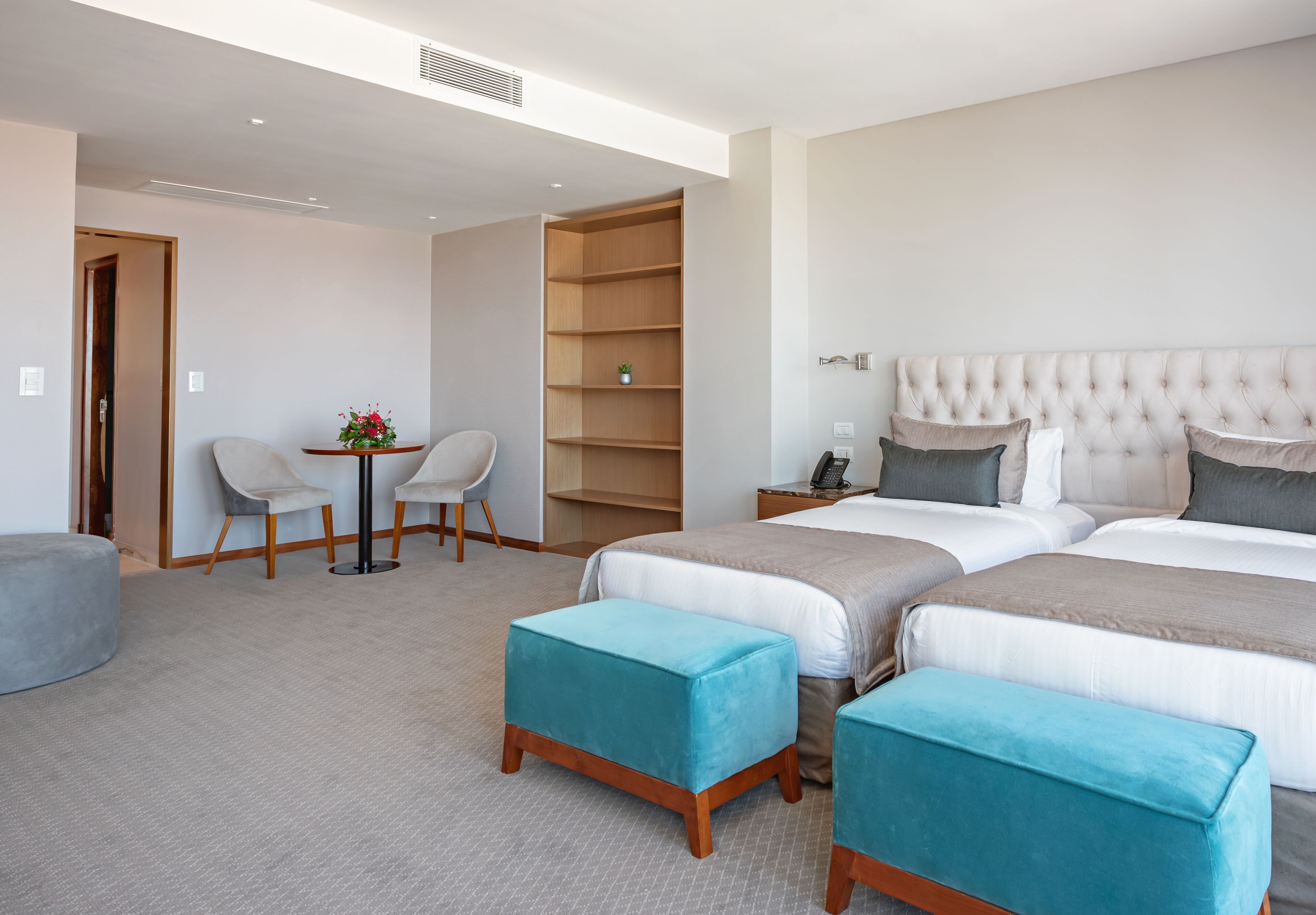 A hotel room with two beds, a seating area with chairs, a small table with a plant, and a bookshelf.
