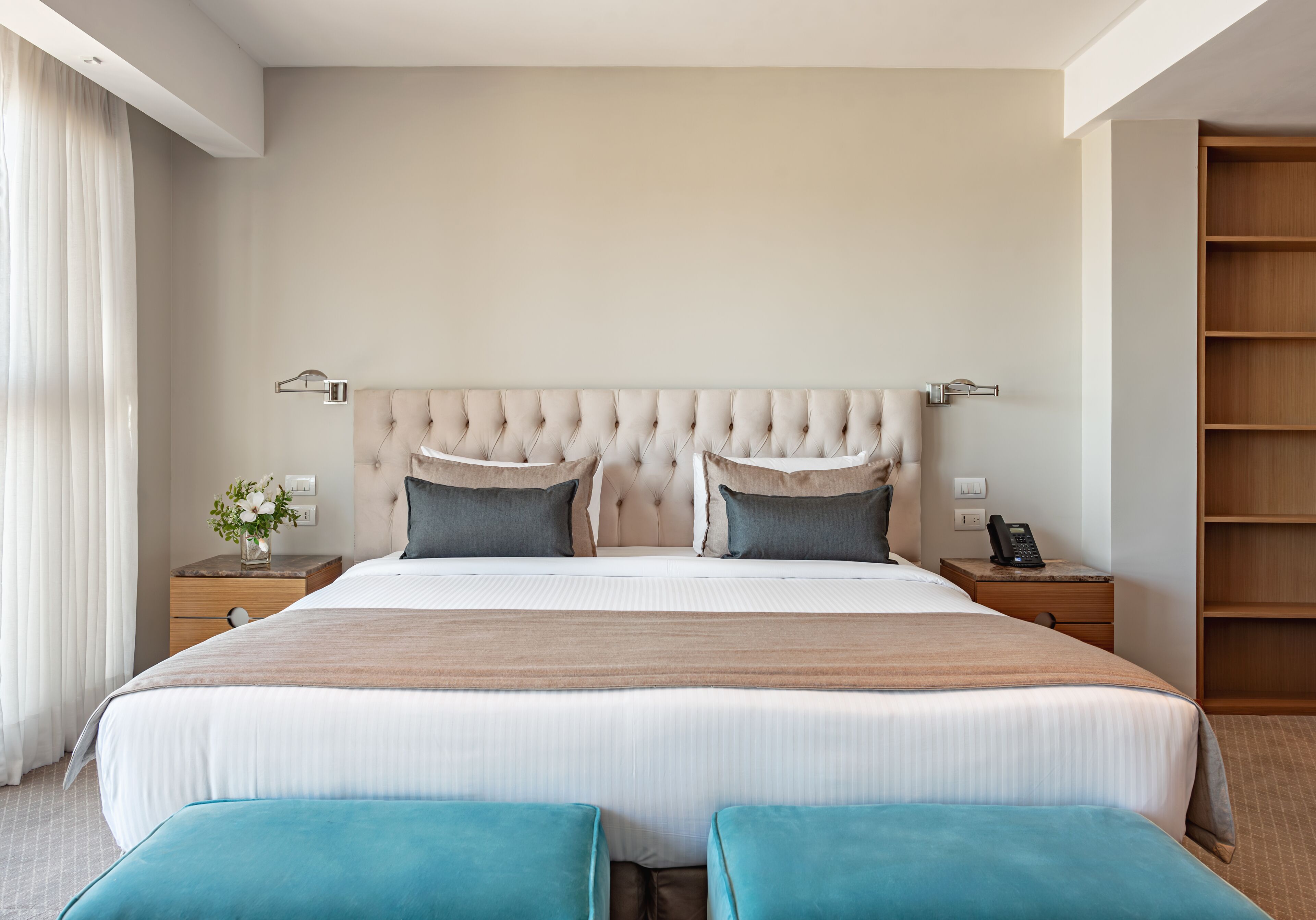 Une chambre d’hôtel moderne avec un grand lit, deux tables de chevet, une petite table, une armoire en bois et une fenêtre avec des rideaux.