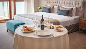 Mesa de jantar bem posta com pratos de comida, copos de vinho e uma garrafa de vinho em quarto de hotel com cama grande e vista para o mar.