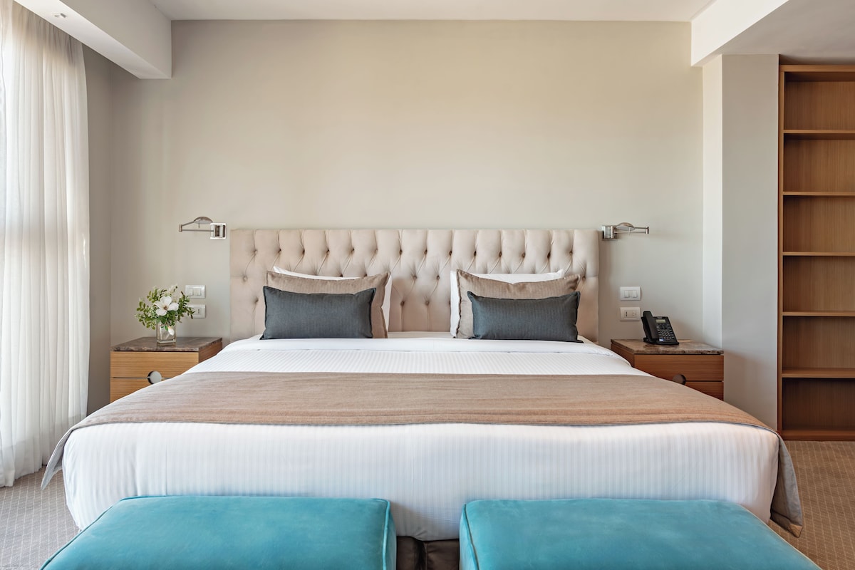 Habitación de hotel moderna con una cama grande, dos mesitas de noche, un armario de madera y una ventana con cortinas.