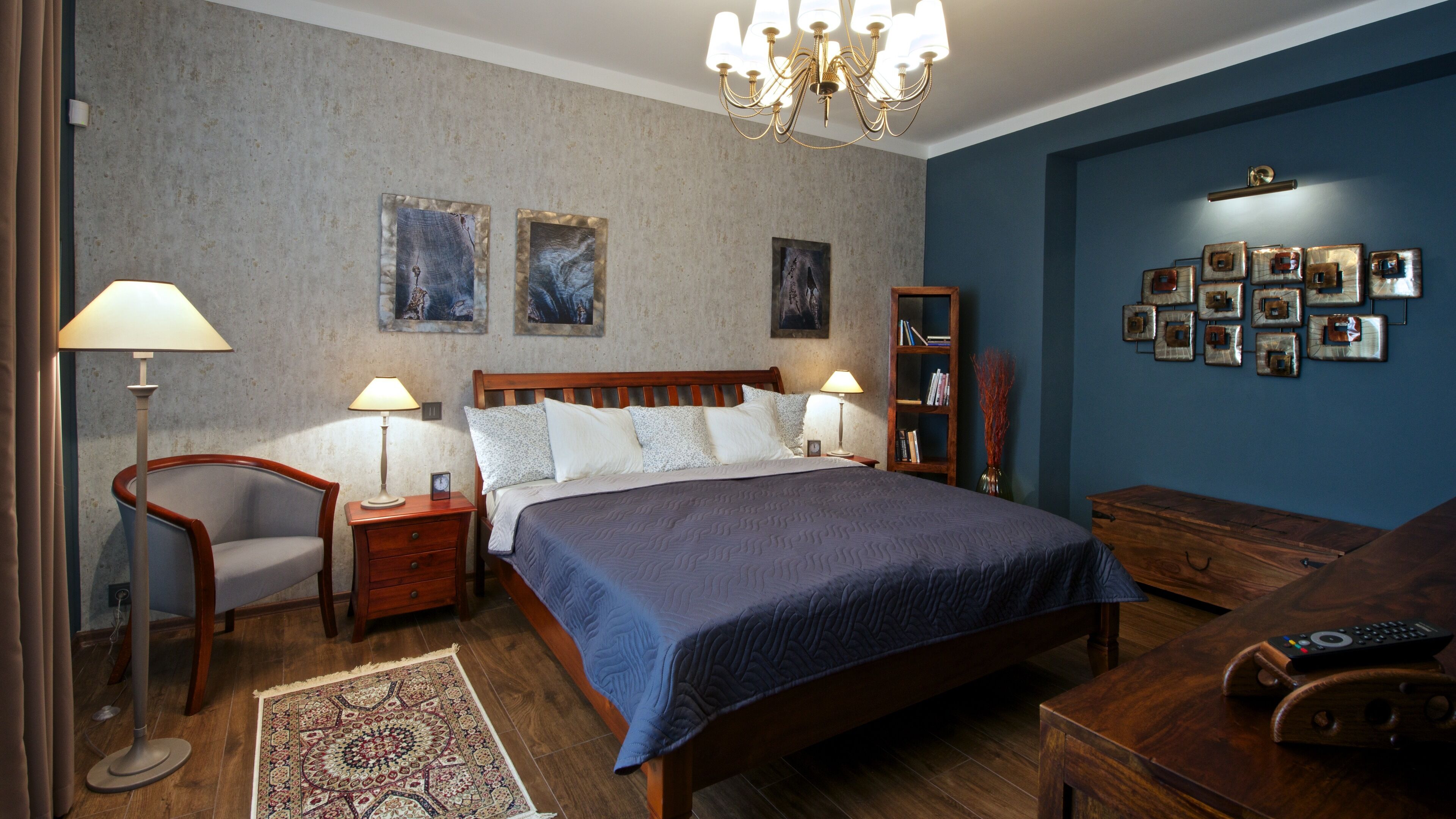 A bedroom with a large bed, two bedside tables with lamps, a wooden nightstand, a chair, a bookshelf, and a wall with framed pictures.
