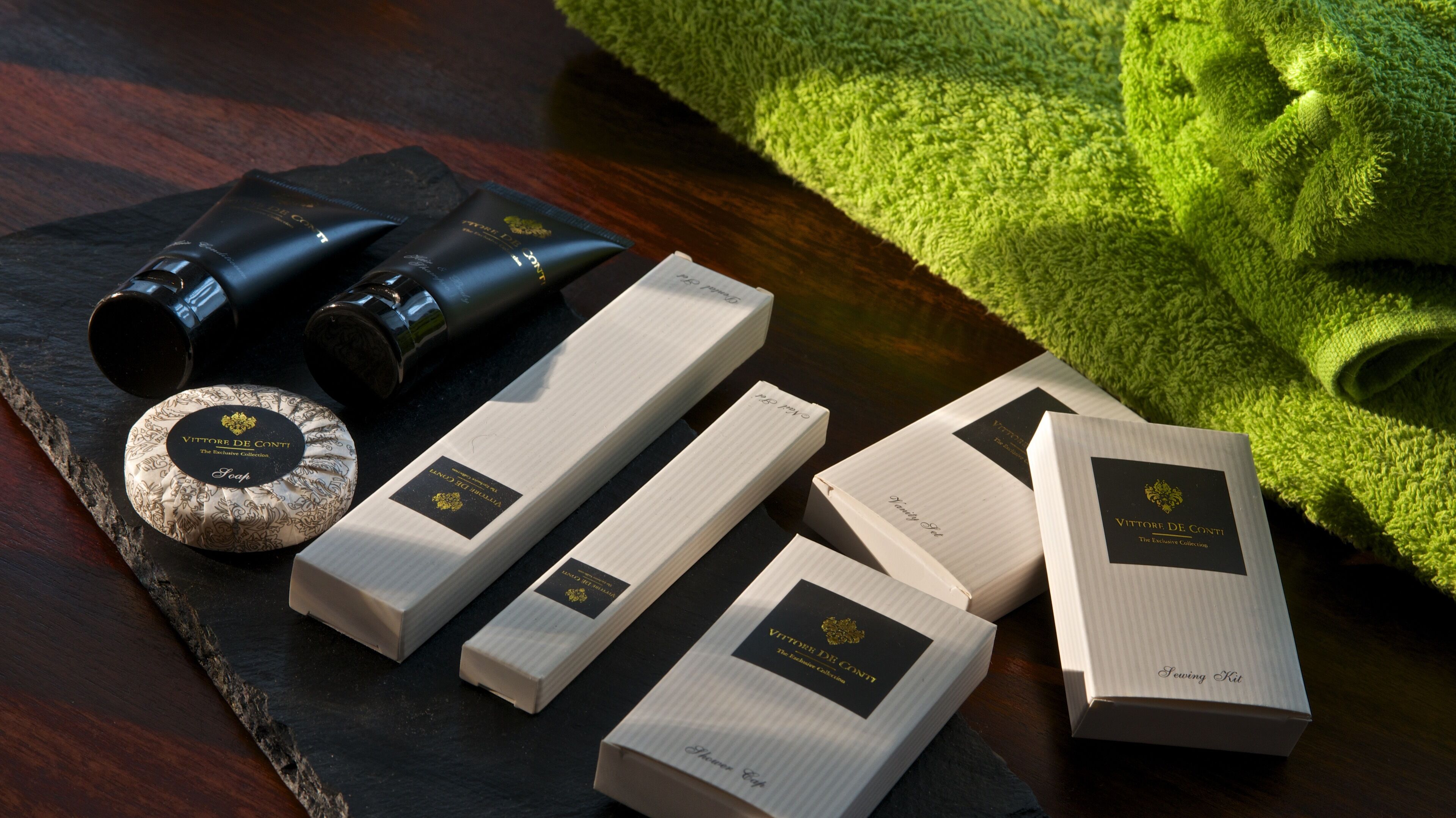 A collection of toiletry items including a soap bar, a sewing kit, and a bottle of lotion, arranged on a dark slate surface with a green towel in the background.