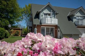 A two-story house with a balcony, a garage, and a well-maintained garden with pink flowers. - Meer und Mehr, Butendieksweg 90c OG, 27476 Cuxhaven- Sahlenburg (Cuxhaven- Sahlenburg)