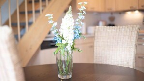 A clear glass vase with white and blue flowers on a wooden table in a room with a staircase and wicker furniture. - Ebbe und Flut, Hannoversche Straße 17 OG, 27476 Cuxhaven- Sahlenburg (Cuxhaven- Sahlenburg)