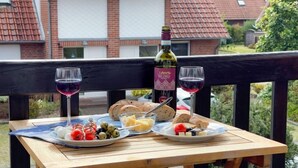 A wooden table with a meal setup including bread, tomatoes, olives, and cheese, accompanied by two glasses of red wine, on a balcony with a house in the background. - Ebbe und Flut, Hannoversche Straße 17 OG, 27476 Cuxhaven- Sahlenburg (Cuxhaven- Sahlenburg)
