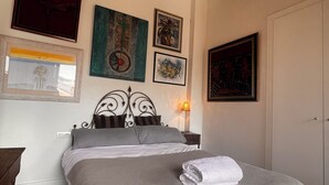 Una cama bien hecha con una colcha gris y blanca, una almohada blanca y una toalla blanca enrollada. Cuatro obras de arte enmarcadas adornan la pared sobre la cama.