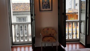 Una habitación con una silla cerca de una ventana, un retrato en un marco en la pared y un balcón con barandilla.