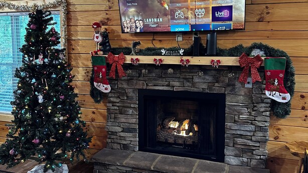 Uma sala de estar aconchegante com lareira de pedra, um pinheiro de Natal e uma televisão de tela plana na parede exibindo a interface de um serviço de streaming.