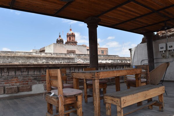 Une terrasse sur le toit, meublée de mobilier en bois, offrant une vue sur un bâtiment historique doté de coupoles et bordée d’un mur de briques.