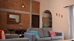 Ein Wohnzimmer mit Backsteinwand, einem Sofa, einem Esstisch und einer durch einen Bogen sichtbaren Küche.