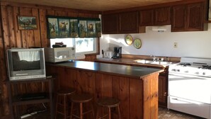 Una cocina con alacenas de madera, un microondas, una estufa, una barra con taburetes y un televisor.