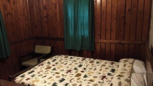 Ein Zimmer mit Holzwänden, einem Bett mit gemustertem Überwurf, einem Stuhl und einem Fenster mit grünen Vorhängen.