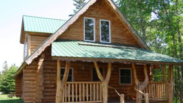 Eine Holzblockhütte mit einem grünen Metalldach, einer Veranda mit Holzgeländern und einer kleinen Treppe zum Eingang.