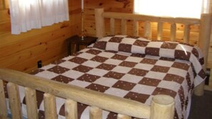 A wooden cabin room with a checkered bedspread, a nightstand, a lamp, and a window with curtains.