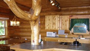 A wooden cabin interior with a central island, wooden cabinets, a microwave, and a window with a green curtain.