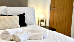 A neatly made bed with white linens and a quilted cover, a bedside table with a lamp, and a wooden door in a hotel room. - *Penthouse Luxury Apartment 2min to Carlisle City (Carlisle)