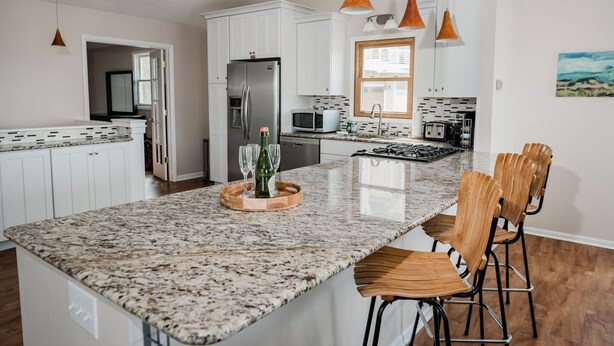 A modern kitchen with white cabinets, granite countertops, a stainless steel refrigerator, and a central island with bar stools. - 4BDR Blue Haven Cottage Grand Haven 150 (Grand Haven)