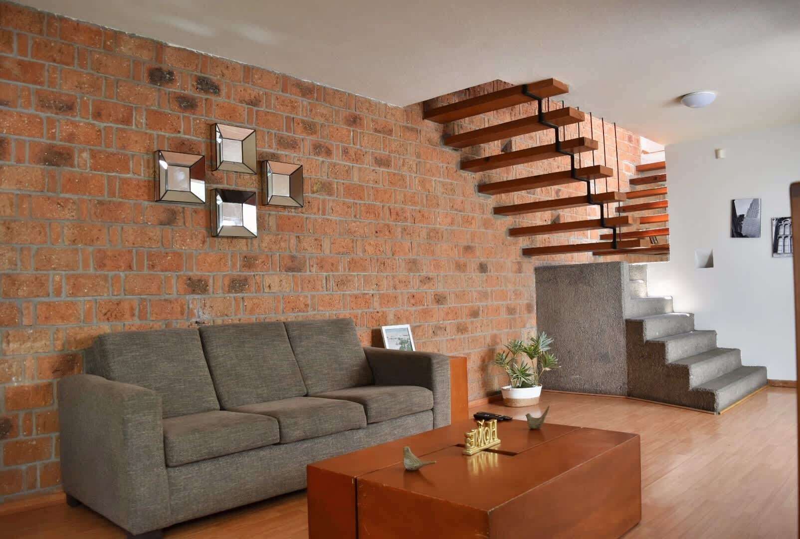 A living room with a brick wall, a grey sofa, a wooden coffee table, and a staircase with a wooden railing.