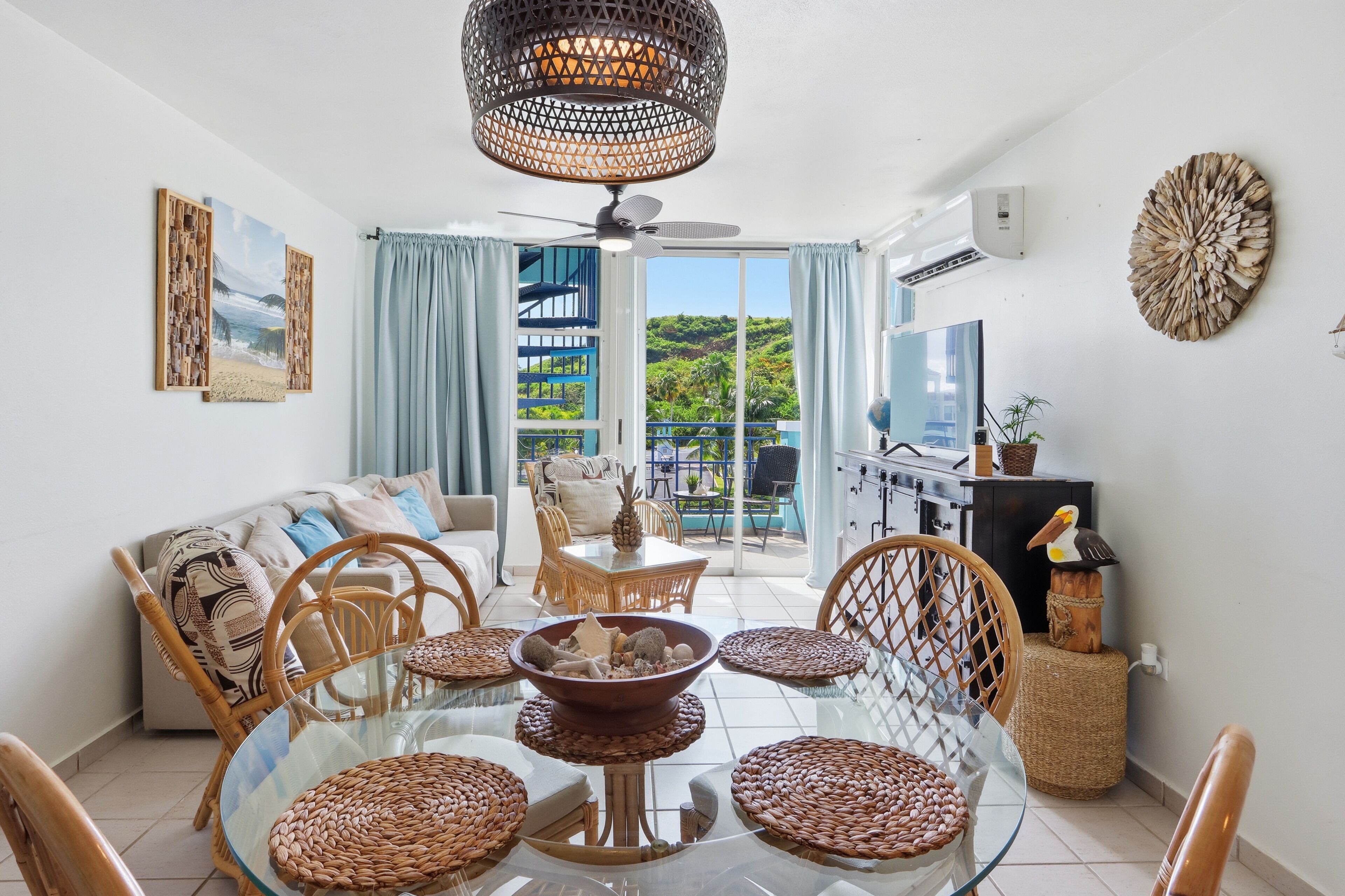 Una sala de estar moderna con una mesa de comedor de vidrio, sillas de mimbre, un sofá y vistas a un jardín exuberante a través de un ventanal corredero.