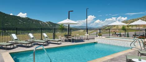 Une piscine entourée d’une clôture, dotée de chaises longues et de parasols, avec une chaîne de montagnes en arrière-plan.