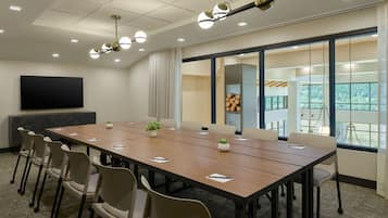 A conference room with a long table, chairs, a flat-screen TV, and a large window with a view of greenery.