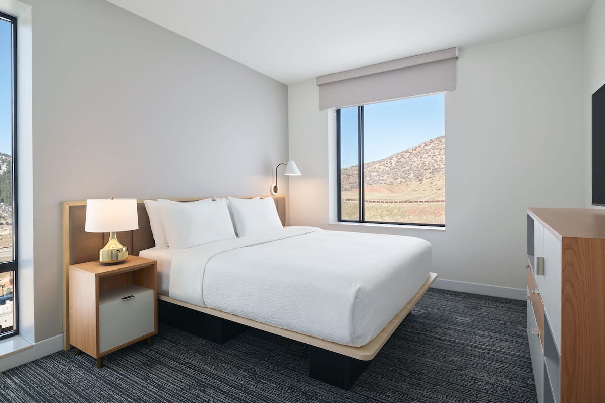 Una habitación de hotel con cama, mesita de noche con lámpara, cómoda y ventana con vista a paisaje desértico.