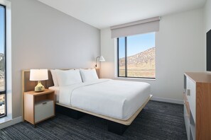 Una habitación de hotel con cama, mesita de noche con lámpara, cómoda y ventana con vista a paisaje desértico.