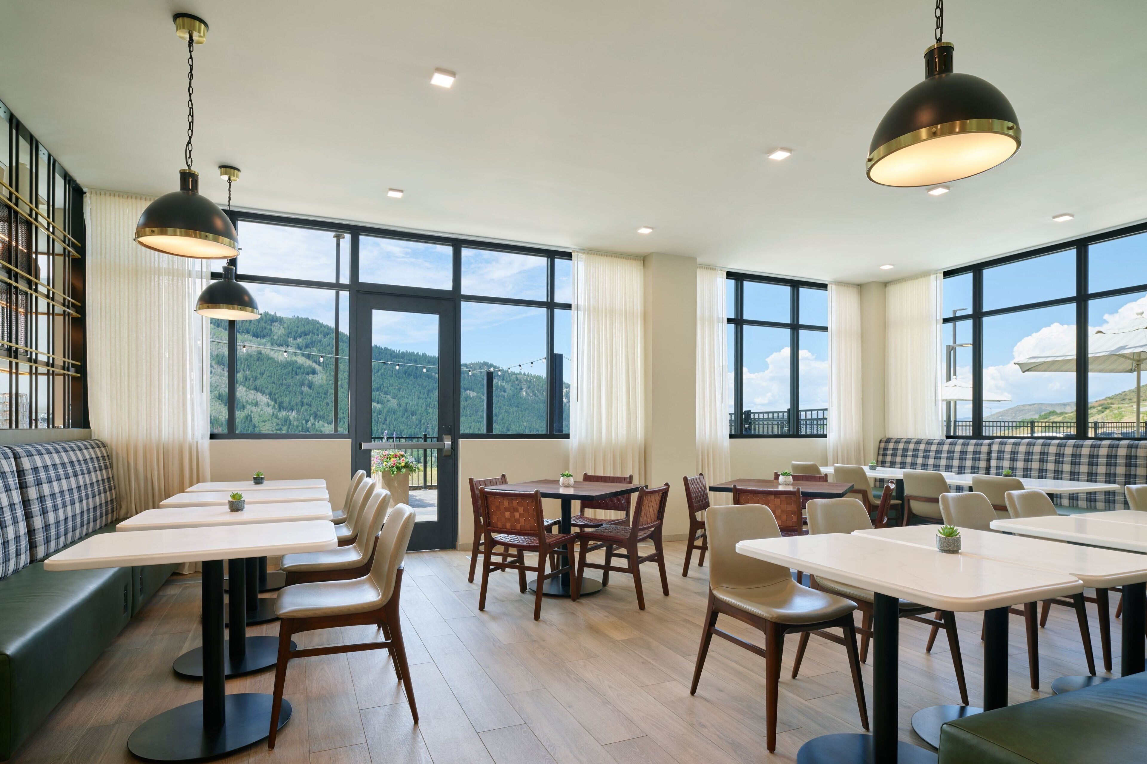 A dining area with multiple tables and chairs, large windows with a view of the outdoors, and hanging pendant lights.