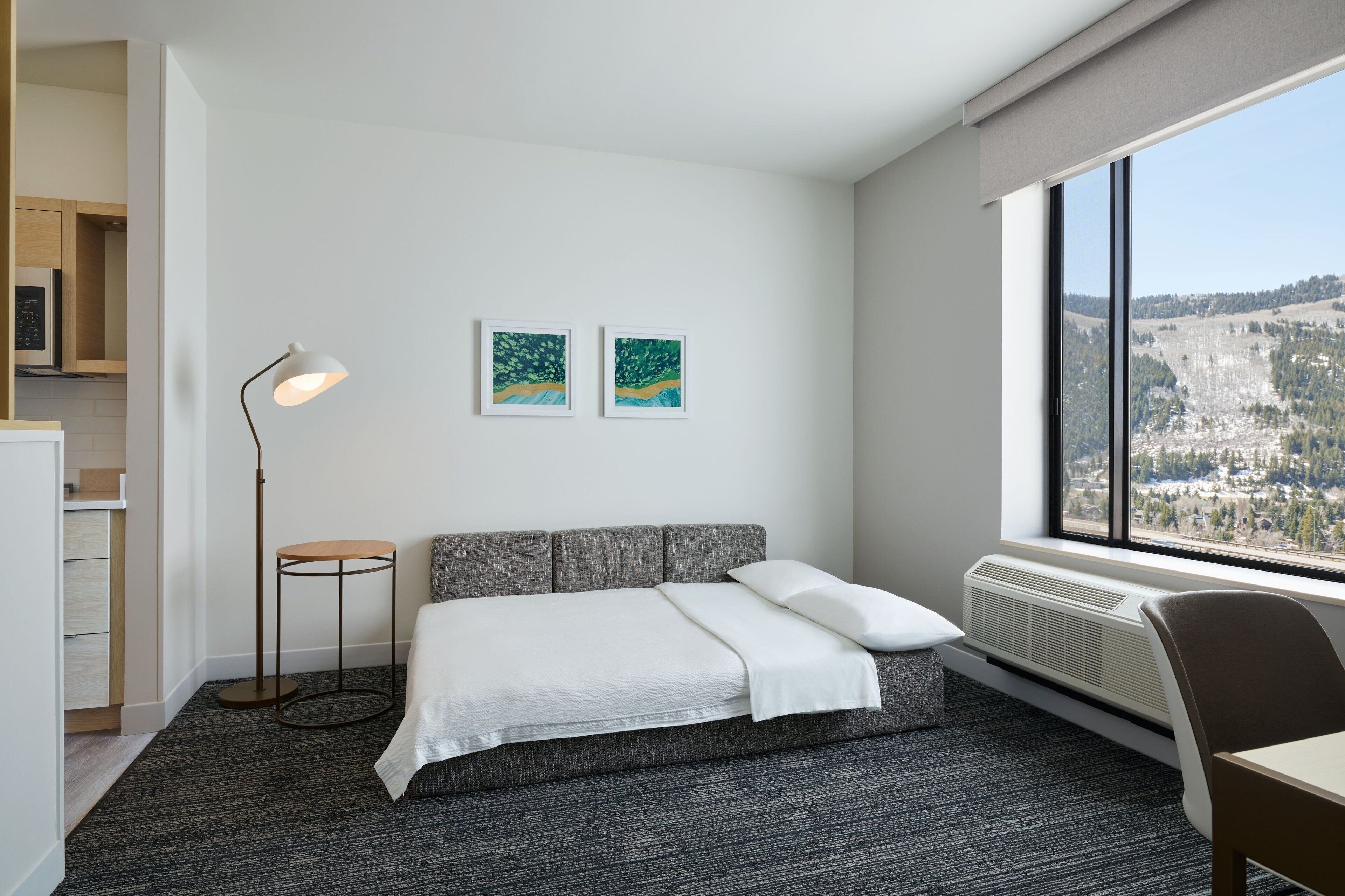 A modern hotel room with a gray sofa bed, a standing floor lamp, two framed artworks on the wall, and a large window offering a view of a mountainous landscape.