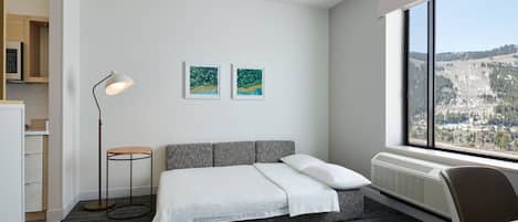 A modern hotel room with a gray sofa bed, a standing floor lamp, two framed artworks on the wall, and a large window offering a view of a mountainous landscape.