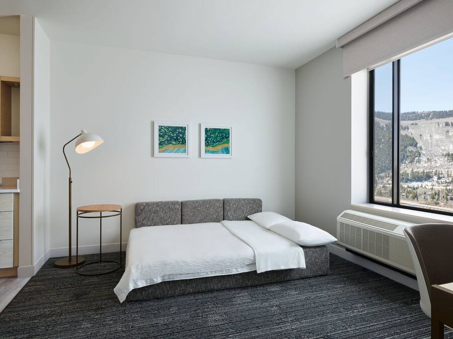 Ein modernes Hotelzimmer mit einer grauen Schlafcouch, einer stehenden Stehlampe, zwei gerahmten Kunstwerken an der Wand und einem großen Fenster mit Blick auf eine gebirgige Landschaft.