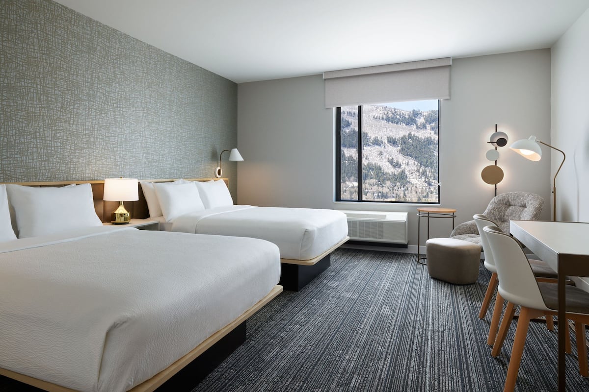 Habitación de hotel con dos camas, un escritorio, una silla, una lámpara y vistas a una montaña nevada.