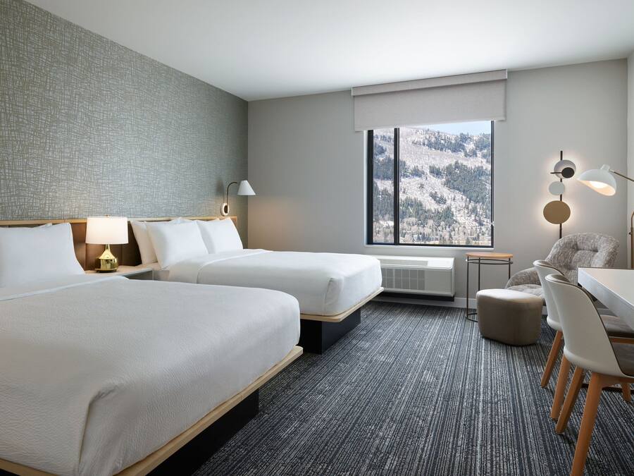 Een hotelkamer met twee bedden, een bureau, een stoel, een lamp en uitzicht op een besneeuwde berg.