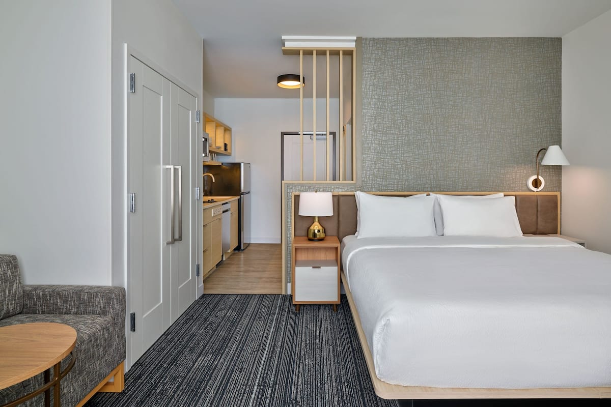 Habitación de hotel con cama, escritorio, silla, mesita de noche, lámpara y kitchenette.