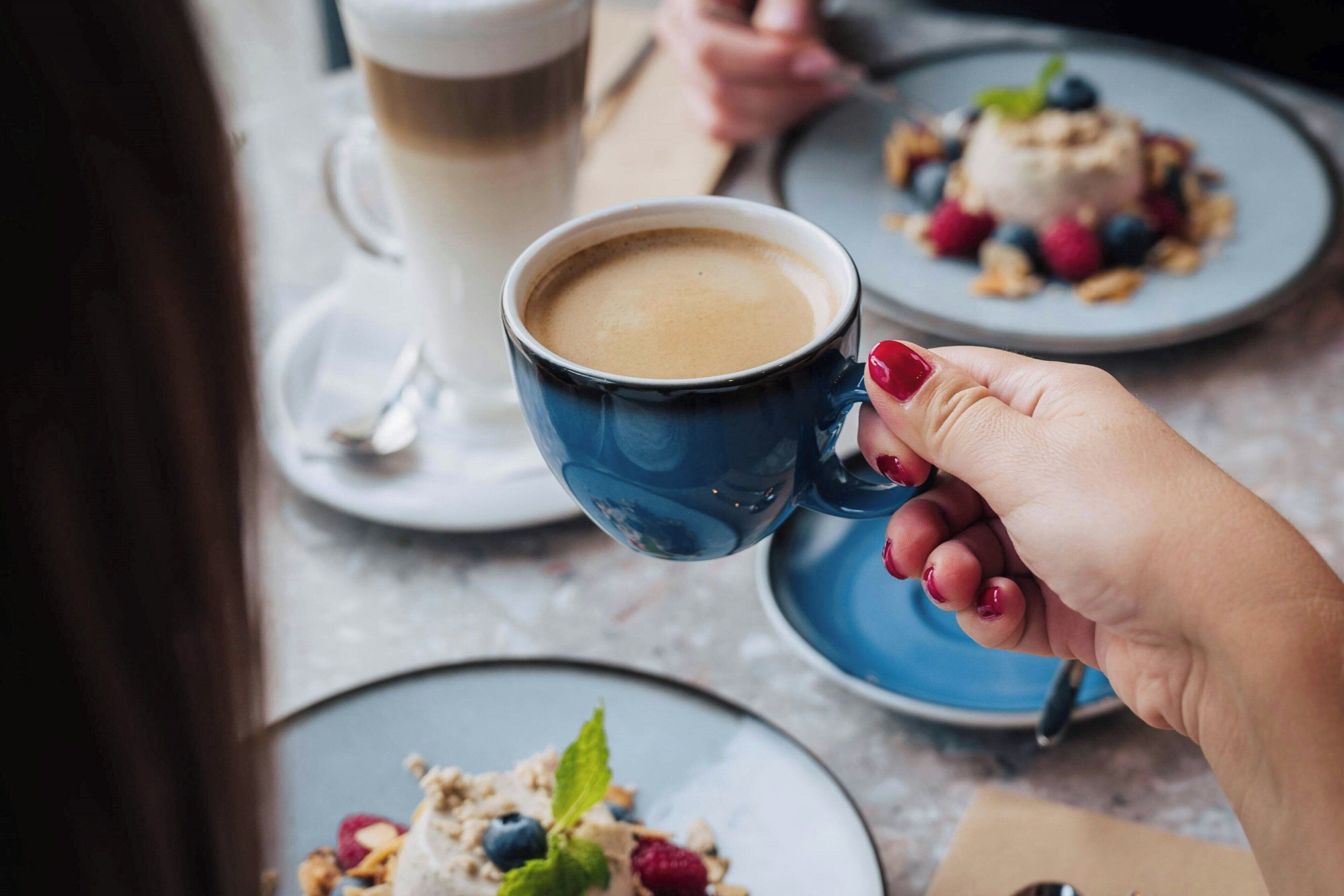 Een persoon houdt een blauwe mok met een drankje vast, een bord met een dessert en een kop koffie staan op een tafel.