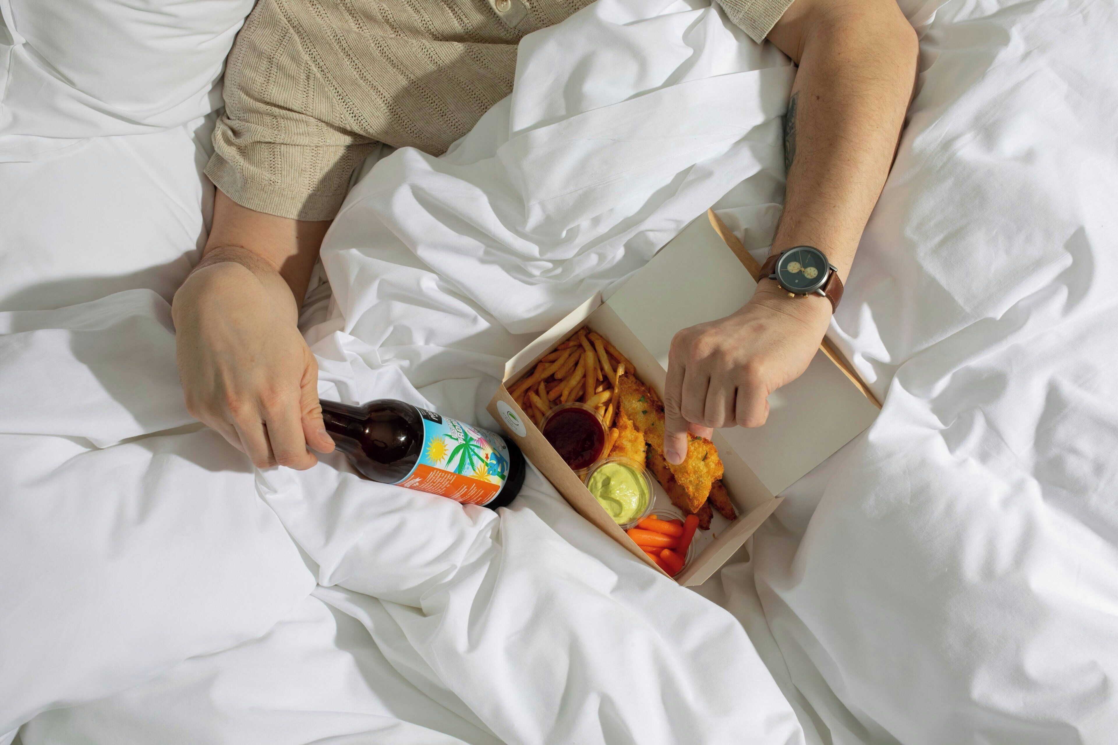 A person lying on a bed with a box of food and a bottle.