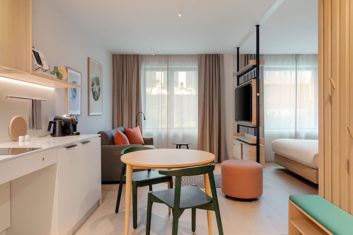 A modern hotel room with a kitchenette, a dining area with a round table and chairs, a sofa, a bed, and a TV.