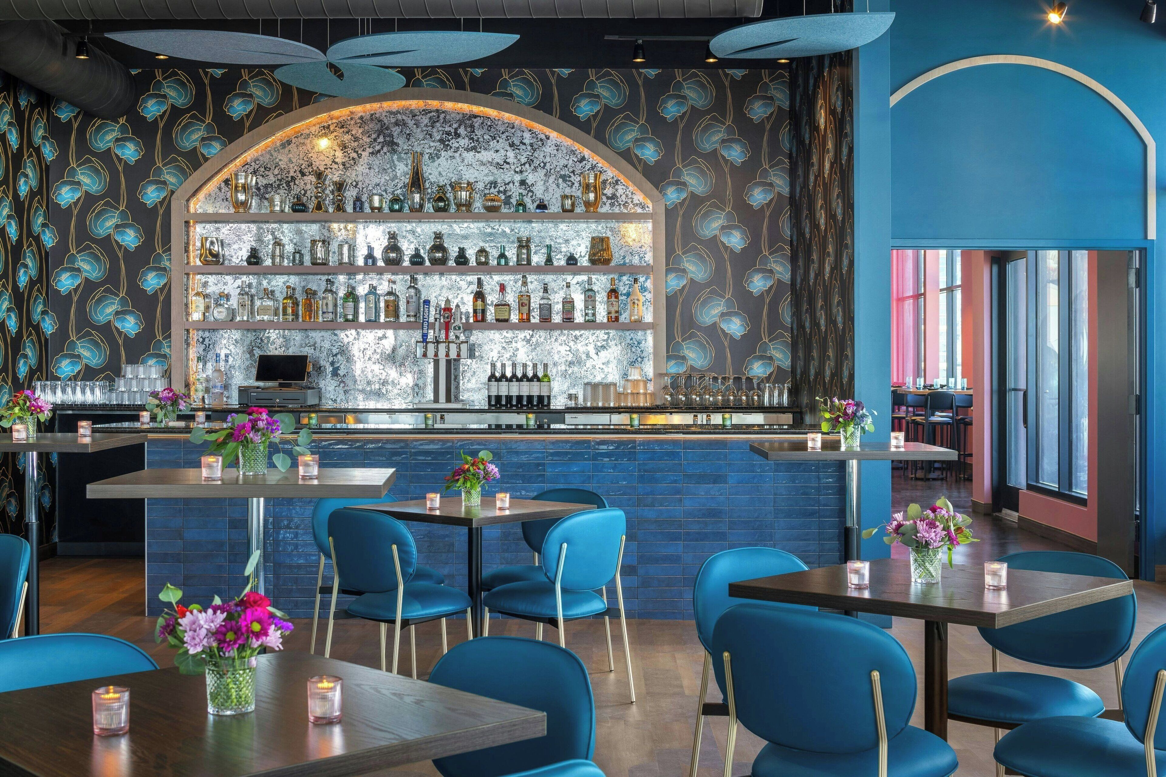 Uma área de bar com esquema de cores azul e dourado, apresentando um balcão, prateleiras com garrafas e cadeiras azuis ao redor das mesas.