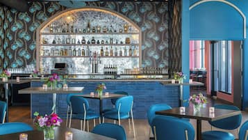 Uma área de bar com esquema de cores azul e dourado, apresentando um balcão, prateleiras com garrafas e cadeiras azuis ao redor das mesas.