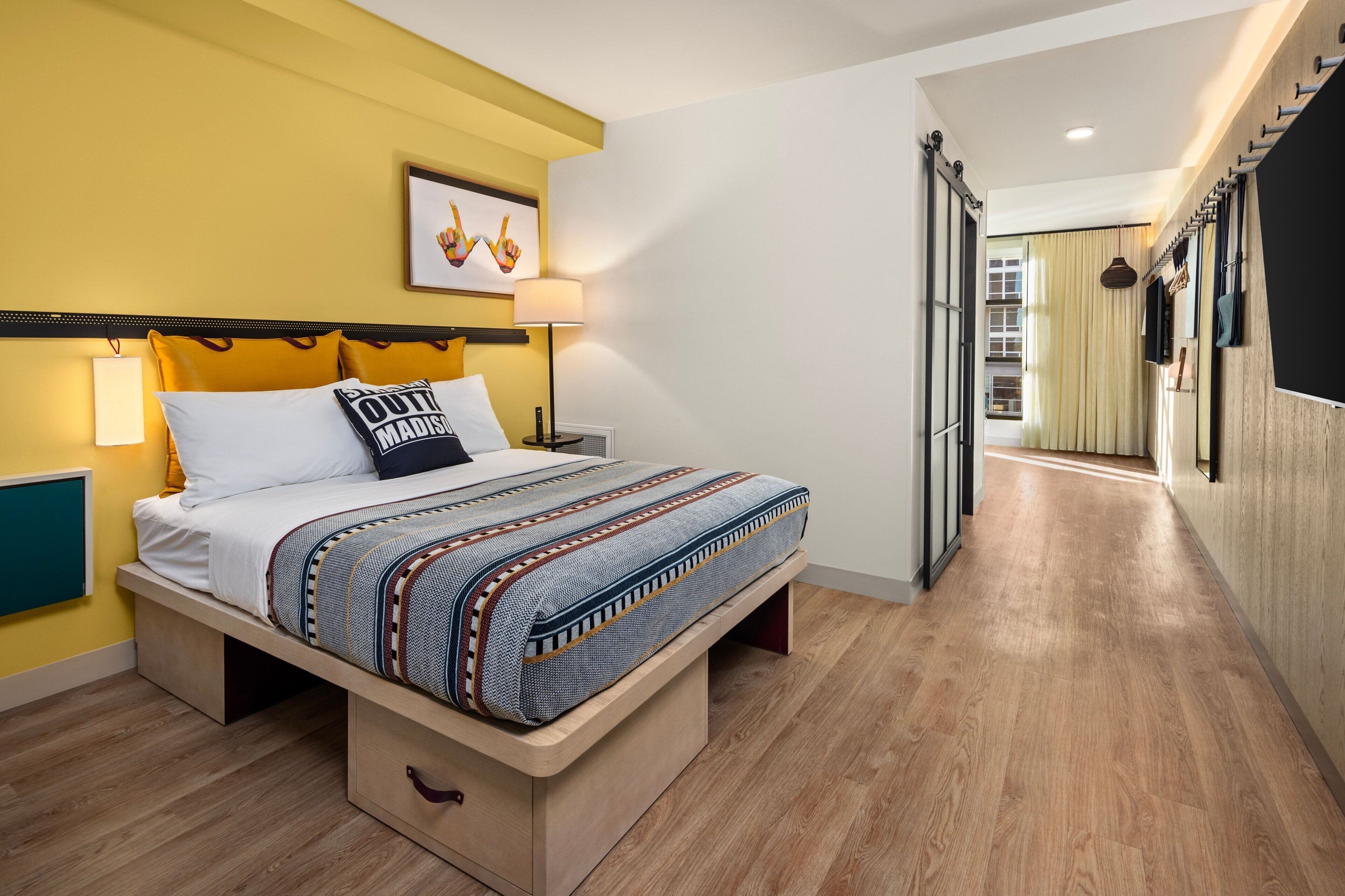 A modern hotel room with a bed, bedside lamps, and a view into the hallway.