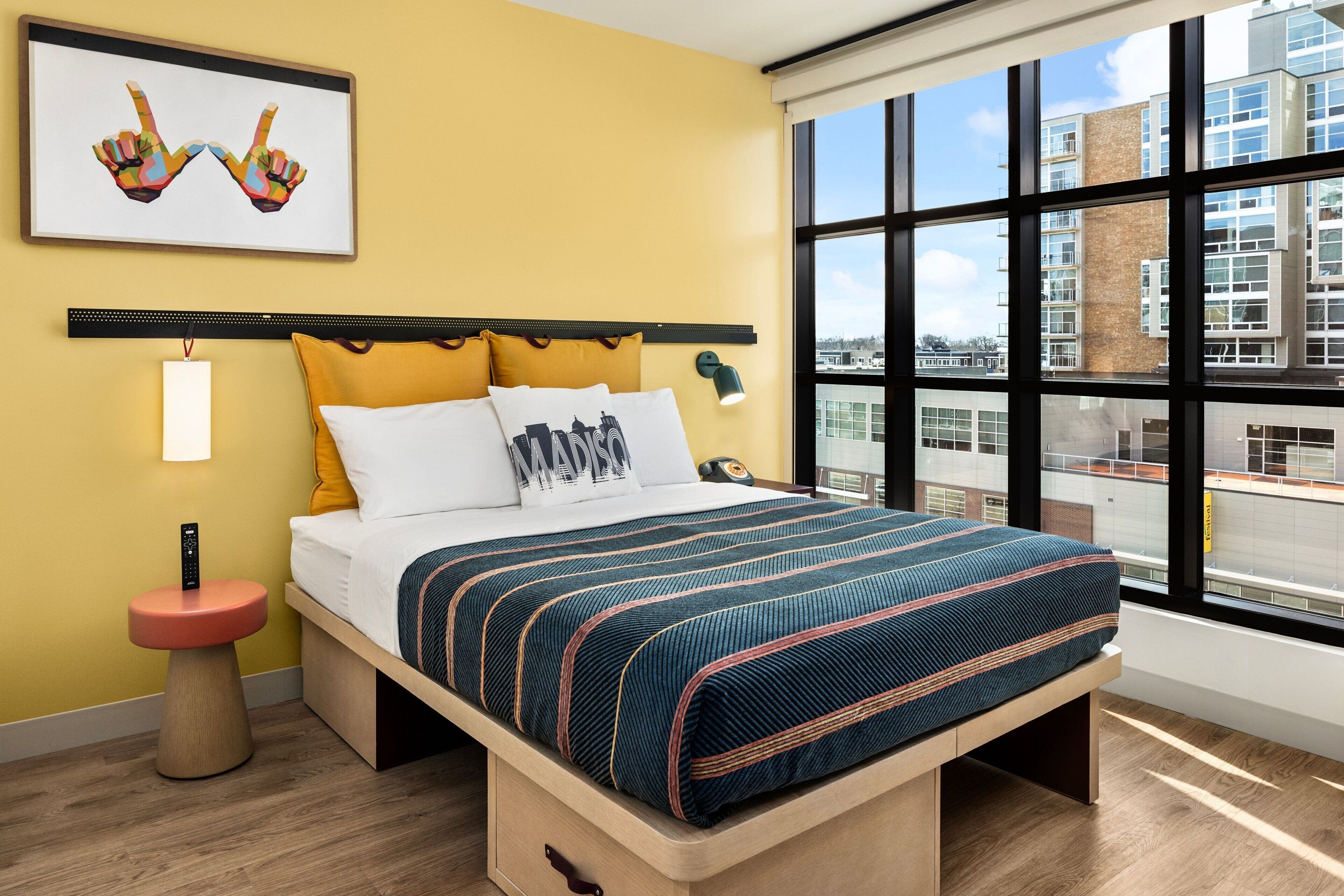 A modern hotel room with a large bed, a bedside table with a remote control, a wall-mounted lamp, and a window with a city view.