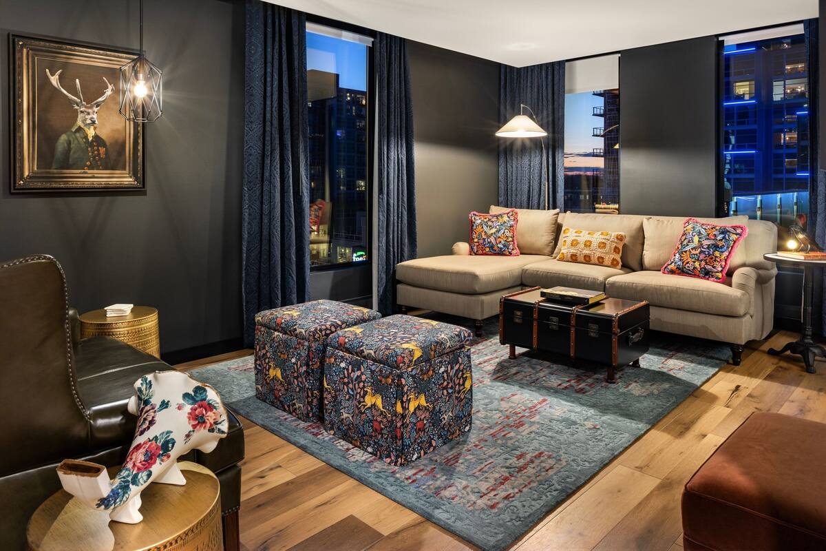 A modern living room with a sectional sofa, patterned ottomans, a framed deer artwork, and a city view through large windows.