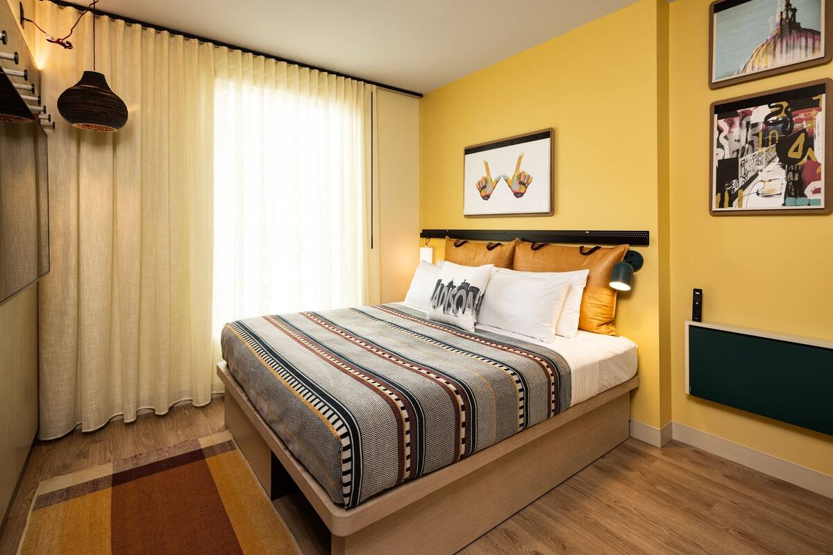 A hotel room with a bed, curtains, a wall-mounted heater, and artwork.